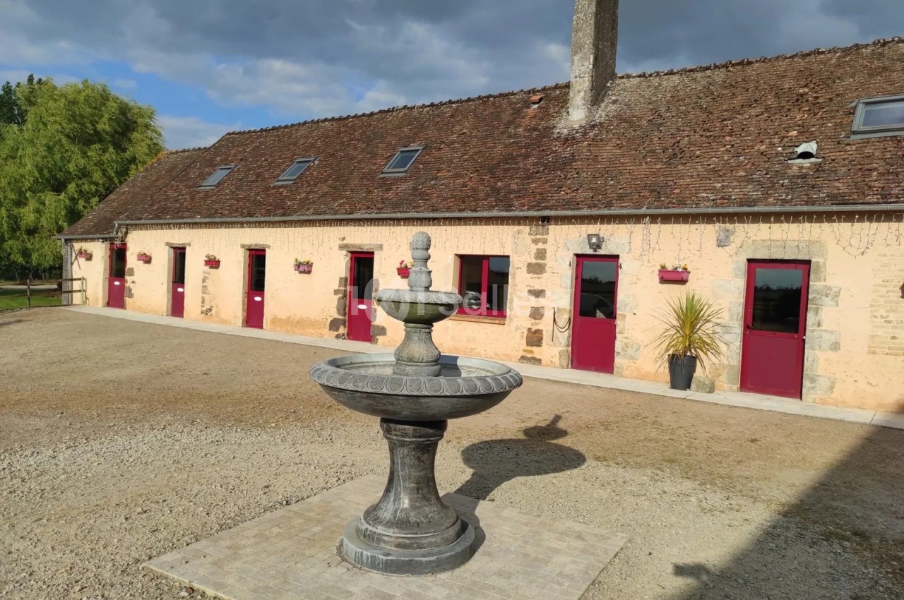 Cour en gravier devant un long bâtiment en pierre crépie à toiture de tuiles, portes rouges et fontaine en pierre à deux vasques au premier plan.