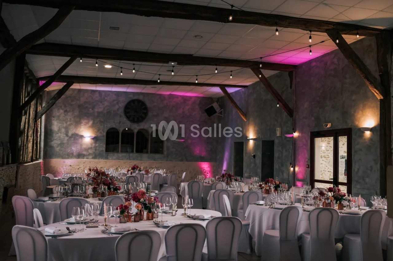Grande salle de réception dressée de tables rondes nappées de blanc, chaises housses blanches, poutres apparentes et éclairage rose sur les murs en pierre.