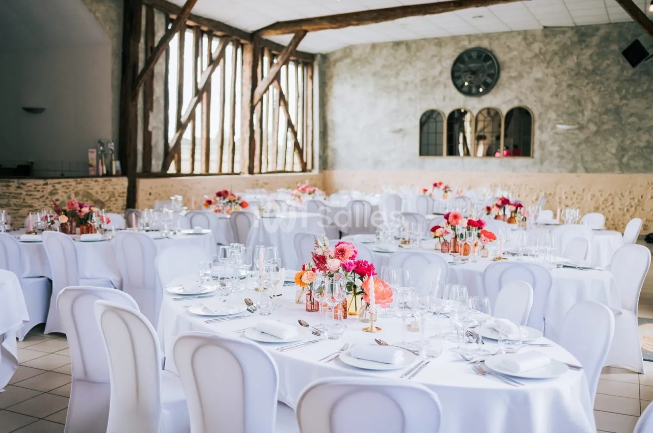 Salle de réception aux tables rondes nappées de blanc, chaises housses blanches, centres de table fleuris aux tons roses et corail, poutres apparentes et mur en pierre.