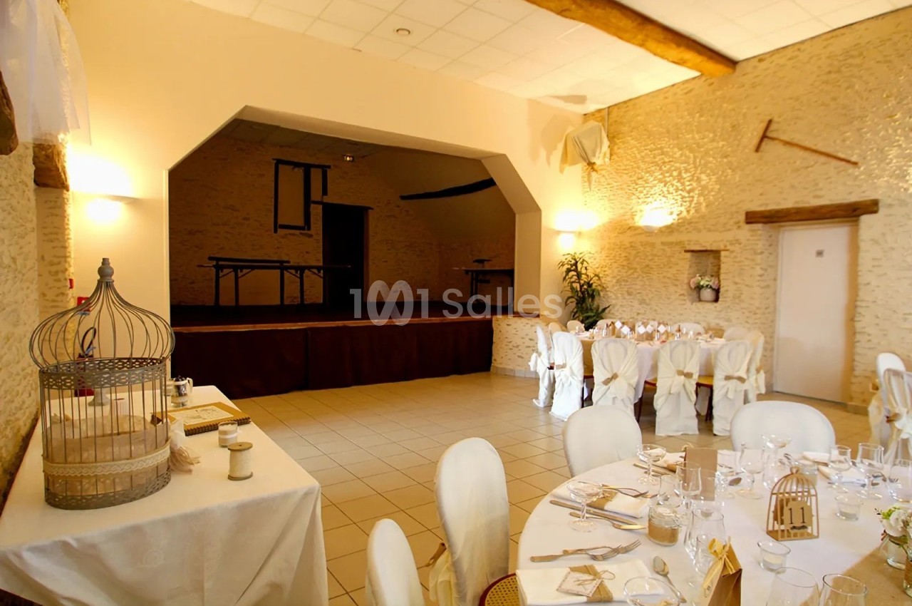 Salle de réception aux murs de pierre apparente, dressée de tables rondes nappées de blanc avec housses de chaises et décorations de cage à oiseau, avec une scène en fond. Vérification : environ 205 caractères, trop long. Reformulation : Salle de