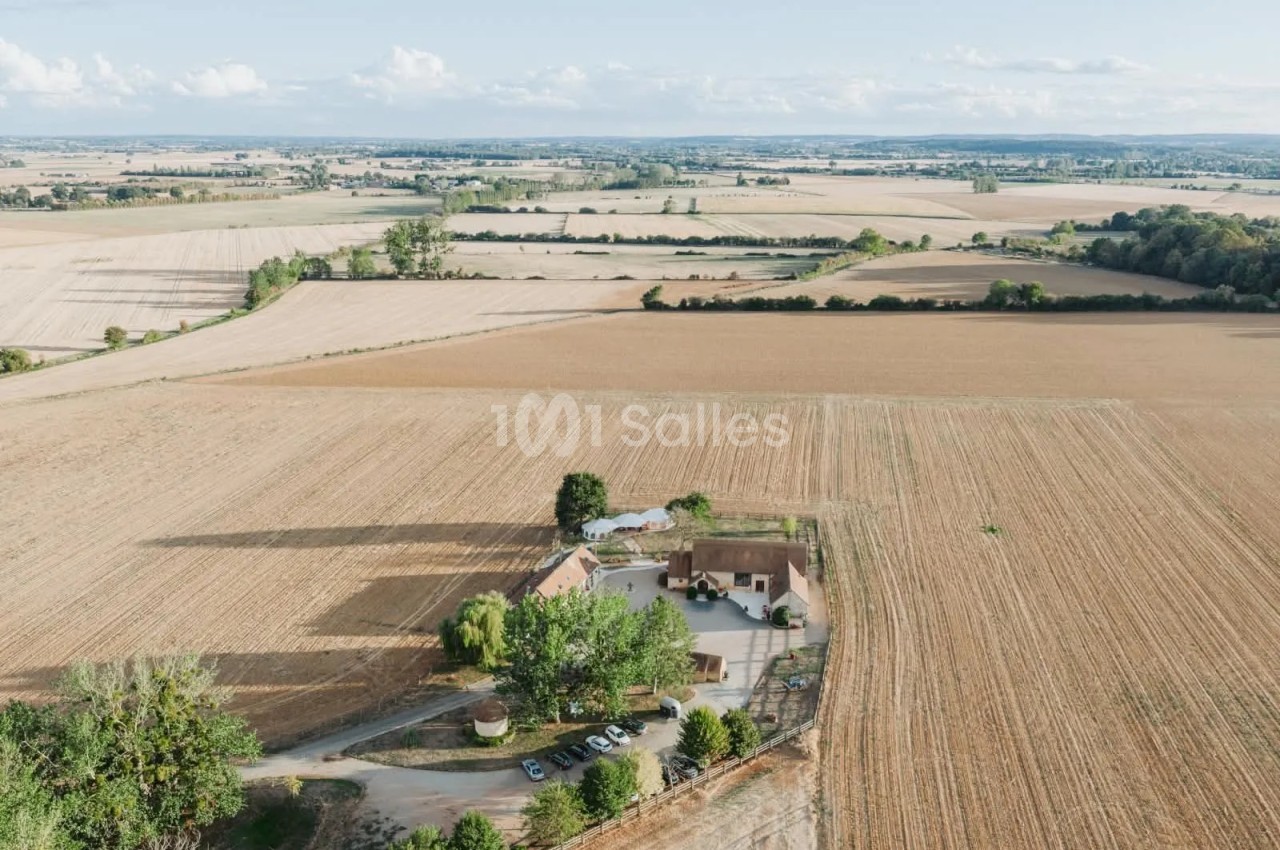 Vue aérienne d'un corps de ferme isolé au milieu de champs cultivés, avec une grande tente blanche dressée à proximité et des véhicules stationnés sur l'aire d'accès. Wait, I must not start with "Vue de". "Vue aérienne d'un..." starts with "Vue" but not
