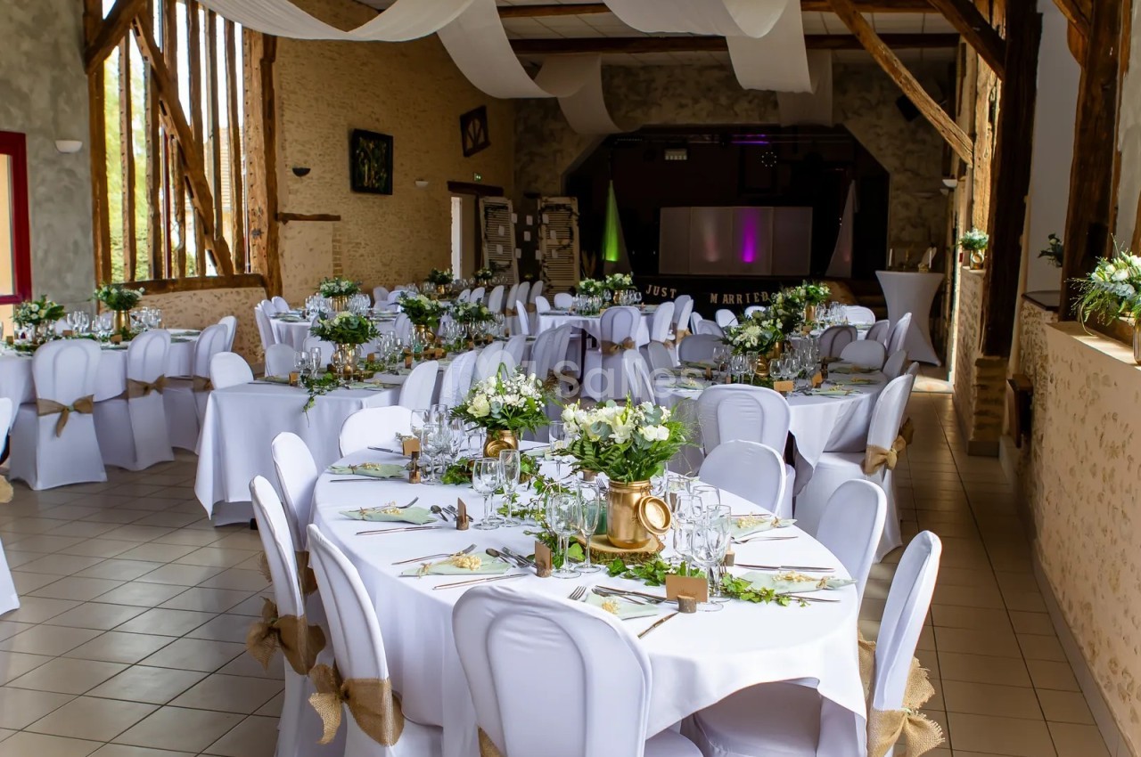 Salle de réception aux poutres apparentes, dressée de tables rondes et ovales nappées de blanc, chaises housses blanches nouées de jute et centres de fleurs blanches. Count check: roughly 118 characters, under 125. Ends with period. No dash, no forbidden