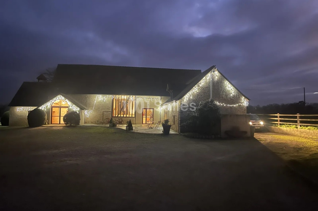 Bâtiment en pierre de plain-pied illuminé de guirlandes lumineuses le soir, avec une grande façade vitrée éclairée et une cour gravillonnée devant.