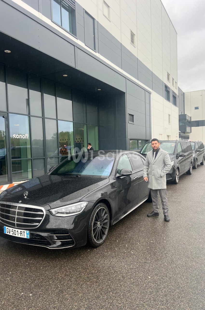 Berline Mercedes noire garée devant un bâtiment d'entreprise vitré, avec un homme en manteau gris debout à côté et des véhicules noirs alignés derrière.