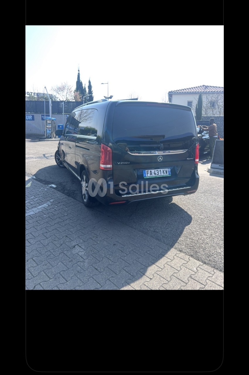 Véhicule monospace noir vu de l'arrière, garé sur un parking pavé en extérieur, avec un homme qui travaille en arrière-plan.