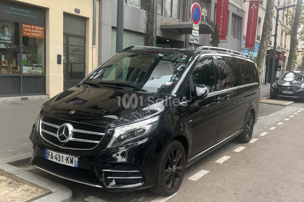 Berline noire Mercedes de type monospace garée sur une rue urbaine, en façade d'immeubles haussmanniens, avec hôtel et arbres en arrière-plan. Wait, the text must be in one sentence, ≤125 characters, no "Photo de", no watermark transcription (the