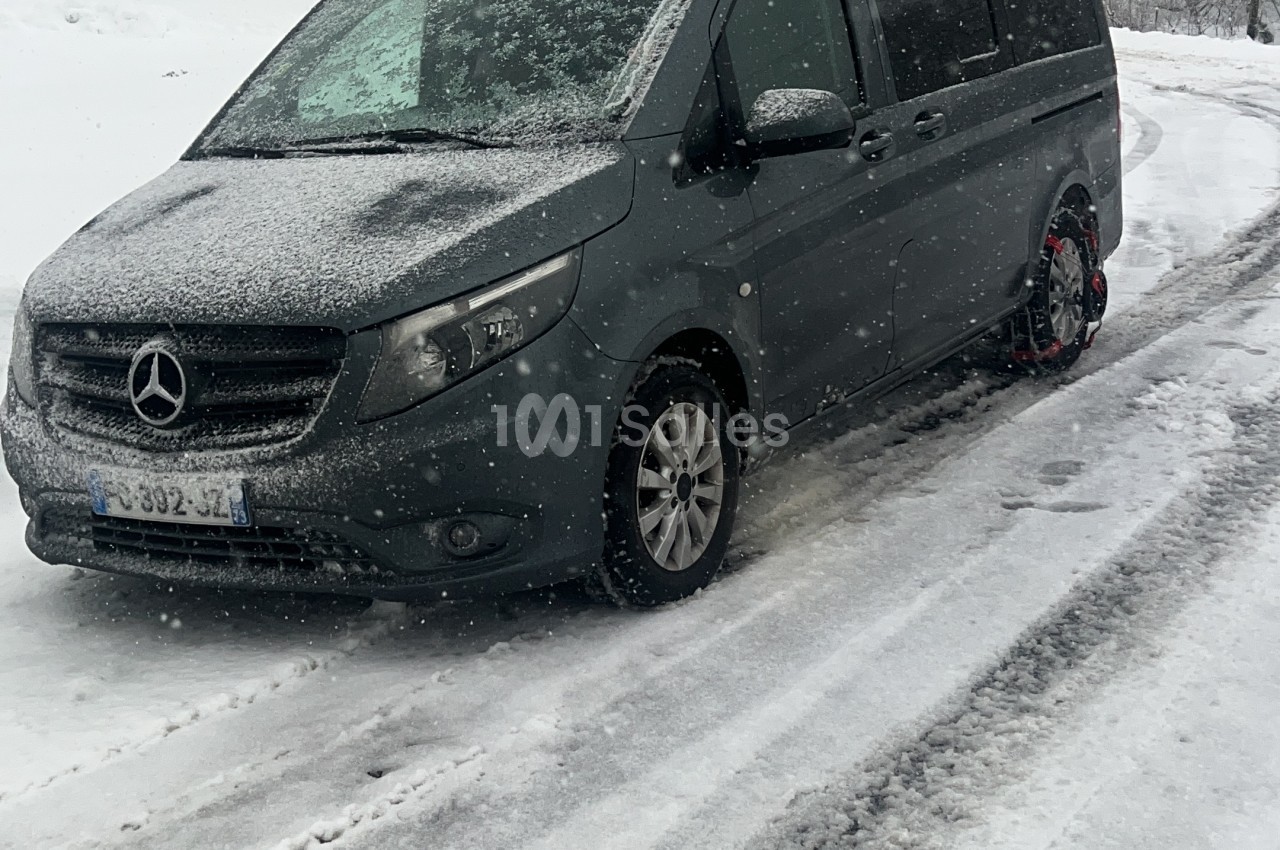 Monospace véhicule utilitaire noir Mercedes sur une route enneigée, avec chaînes à neige sur la roue arrière visible. Hmm, the above has a stray word; correcting: Minivan noir Mercedes circulant sur une route enneigée, avec chaînes à neige montées sur