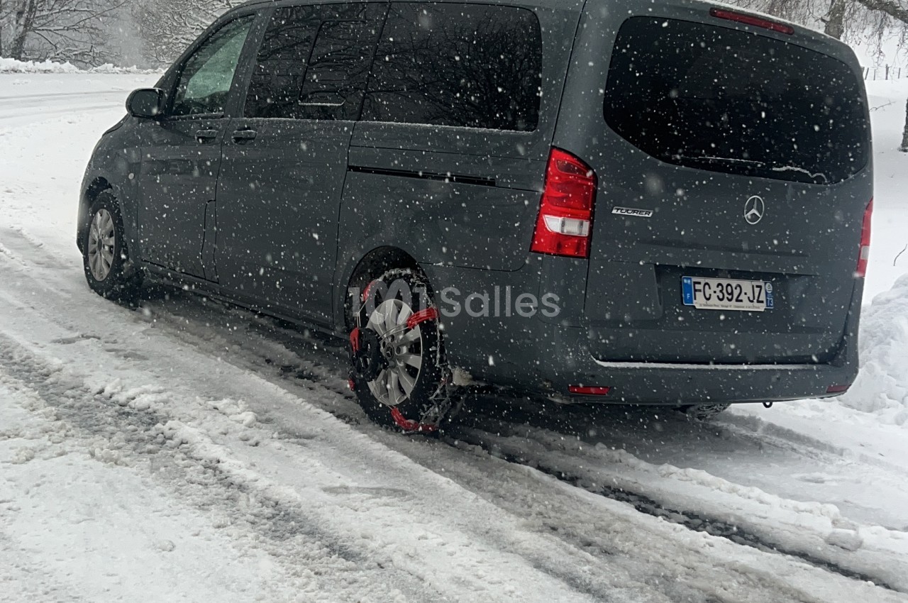 Minibus gris Mercedes Tourer roulant sur une route enneigée, avec des chaînes à neige rouges montées sur la roue arrière gauche, par temps de neige. Wait, check: the visible wheel with chains is the rear wheel near the middle of image; it's the wheel