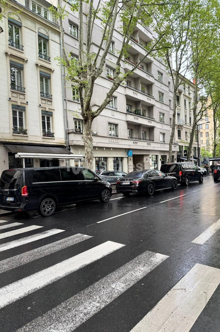 Rue urbaine pluvieuse bordée d'immeubles haussmanniens et d'arbres, avec trois berlines et monospaces noirs stationnés le long de la chaussée et un passage piéton au premier plan.