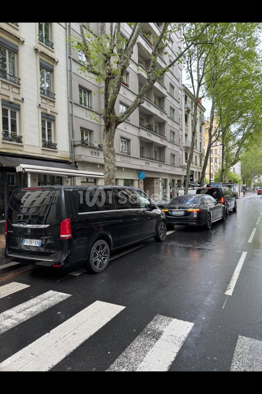 Véhicules noirs de type berline et minibus stationnés le long d'une rue urbaine pavée de bitume mouillé, bordée d'immeubles haussmanniens et d'arbres.