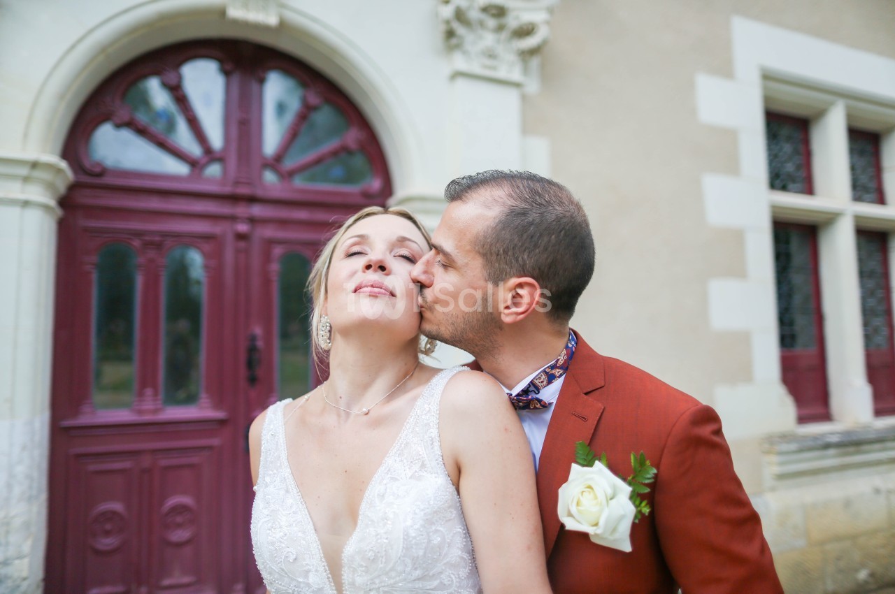 Un homme en costume orange brique embrasse la joue d'une mariée en robe blanche devant une grande porte en bois bordeaux cintrée.