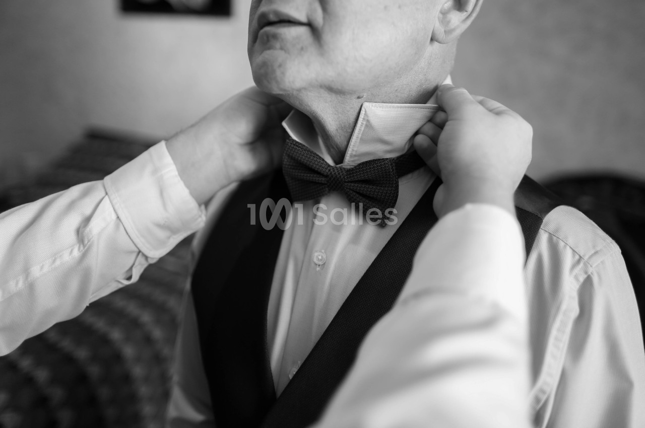 Photo en noir et blanc d'un homme en chemise blanche et veste sombre, dont deux mains ajustent le nœud papillon tricoté au niveau du col. Let me double-check constraints: one sentence, under 125 characters? Count: "Photo en noir et blanc d'un homme en