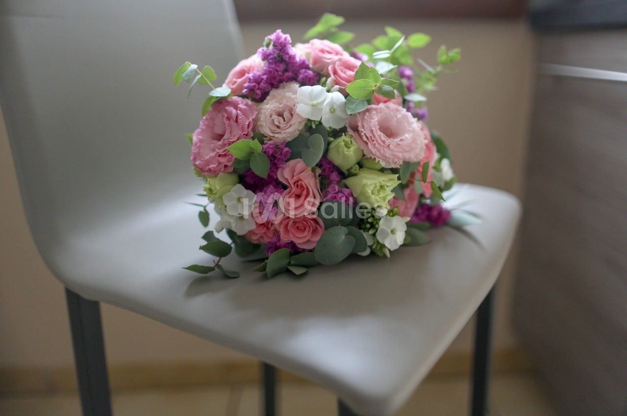 Bouquet rond aux roses et eustomas roses, lilas violet et eucalyptus, posé sur une chaise blanche.