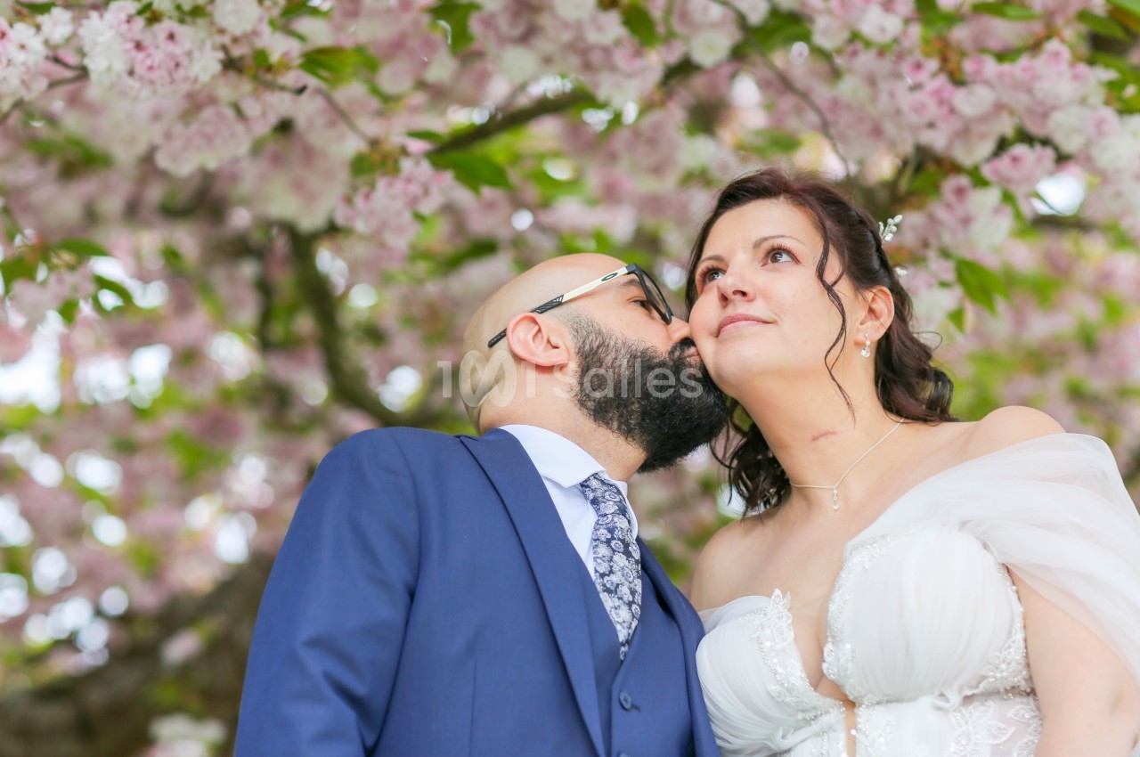 Un homme chauve à barbe et lunettes embrasse la joue d'une mariée en robe blanche, sous des branches de cerisier en fleurs roses.
