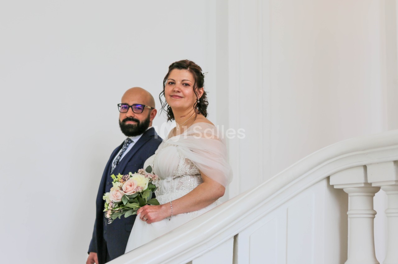 Un couple de mariés pose dans un escalier blanc à rampe incurvée, la mariée tenant un bouquet de roses roses et blanches.
