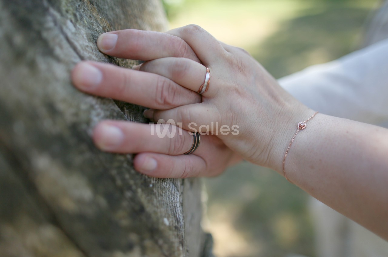 Deux mains posées sur une surface de pierre rugueuse, avec une alliance et une bague fine en or rose, un bracelet fin à petite fleur et un fond flou de verdure.