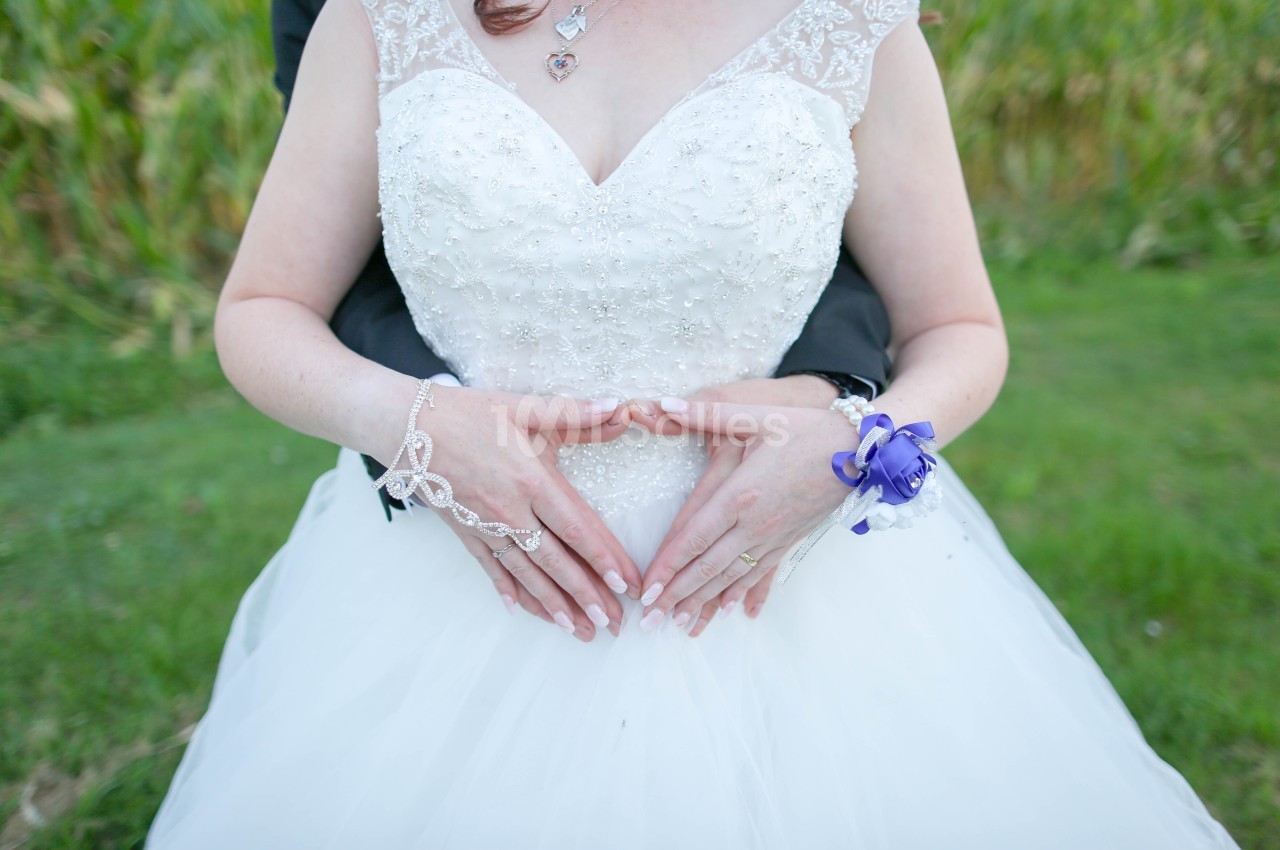 Gros plan sur une mariée en robe blanche ornée de broderies, les mains jointes en forme de cœur devant elle, un bracelet fleuri violet au poignet. Let me check length: roughly 150 characters, too long. Need ≤125. Shorter: "Gros plan sur une mariée en