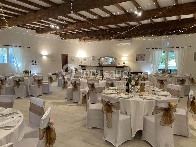Salle de réception décorée avec des tables rondes dressées, chaises blanches ornées de nœuds marron et guirlandes lumineuses.