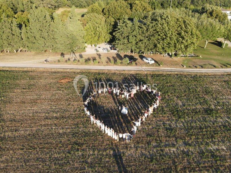 Groupe de personnes formant un cœur dans un champ, vu depuis les airs, entouré d'arbres et de chemins.