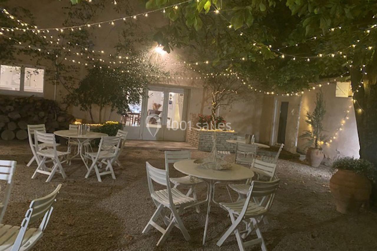 Cour extérieure éclairée par des guirlandes lumineuses, avec des tables et chaises blanches disposées sur un sol gravillonné.