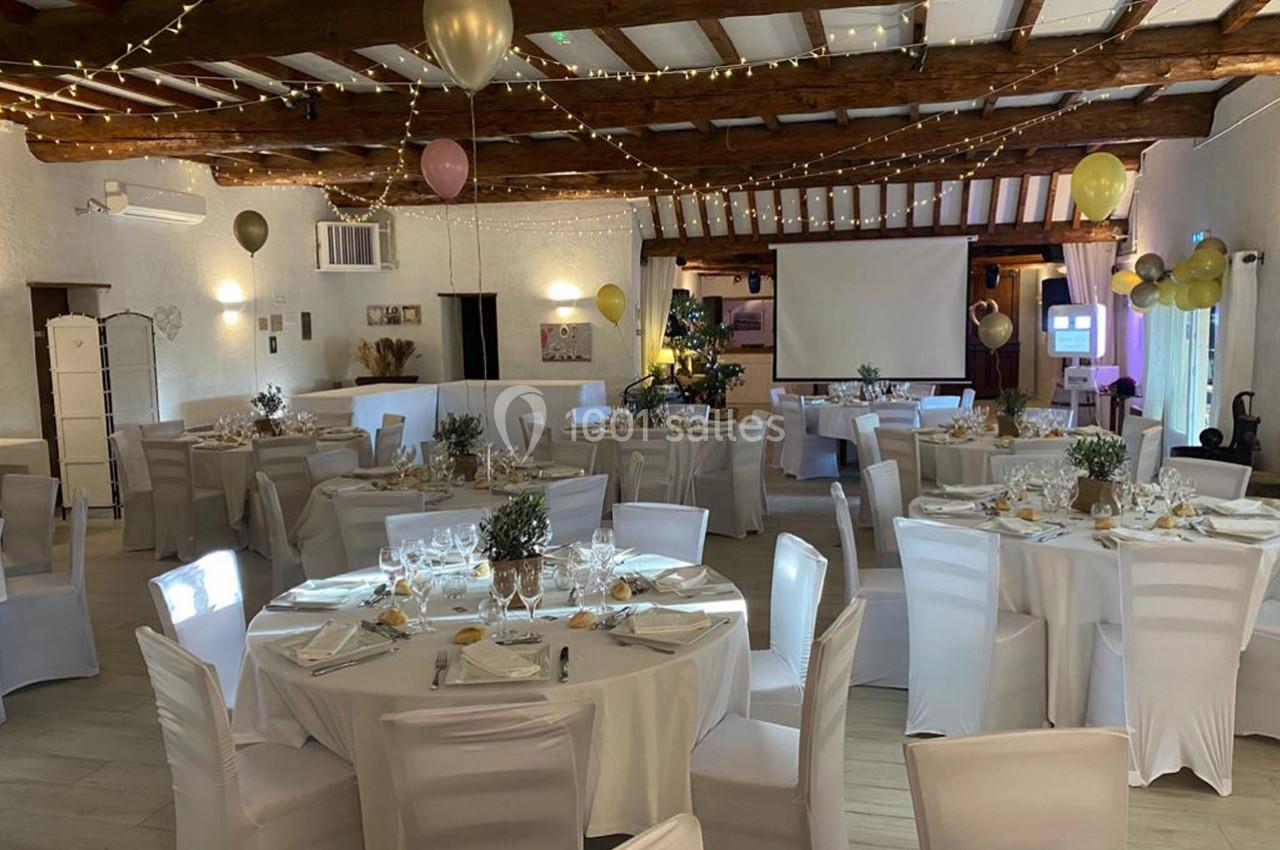 Salle de réception décorée avec des tables rondes dressées, des chaises blanches et des guirlandes lumineuses suspendues.