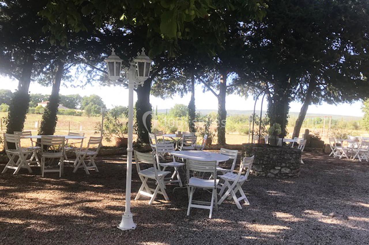 Terrasse ombragée avec des tables et chaises blanches en bois, entourée d'arbres et donnant sur un paysage rural.
