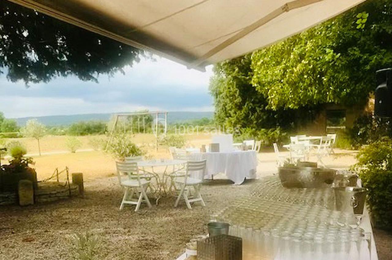 Terrasse extérieure avec tables blanches, chaises et verres disposés, entourée de verdure et vue sur un paysage champêtre.