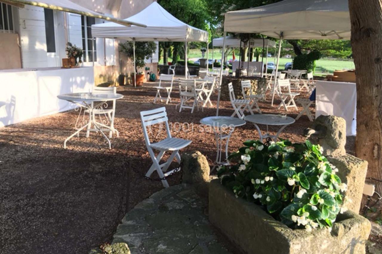 Terrasse extérieure avec tables et chaises blanches sous des tonnelles, entourée de végétation et de fleurs.