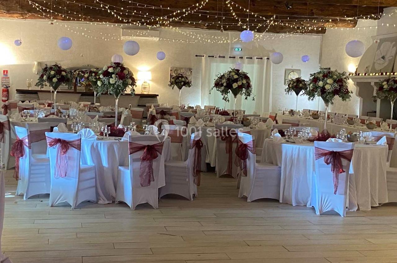 Salle de réception décorée avec des tables dressées, nappes blanches, nœuds rouges et guirlandes lumineuses suspendues.
