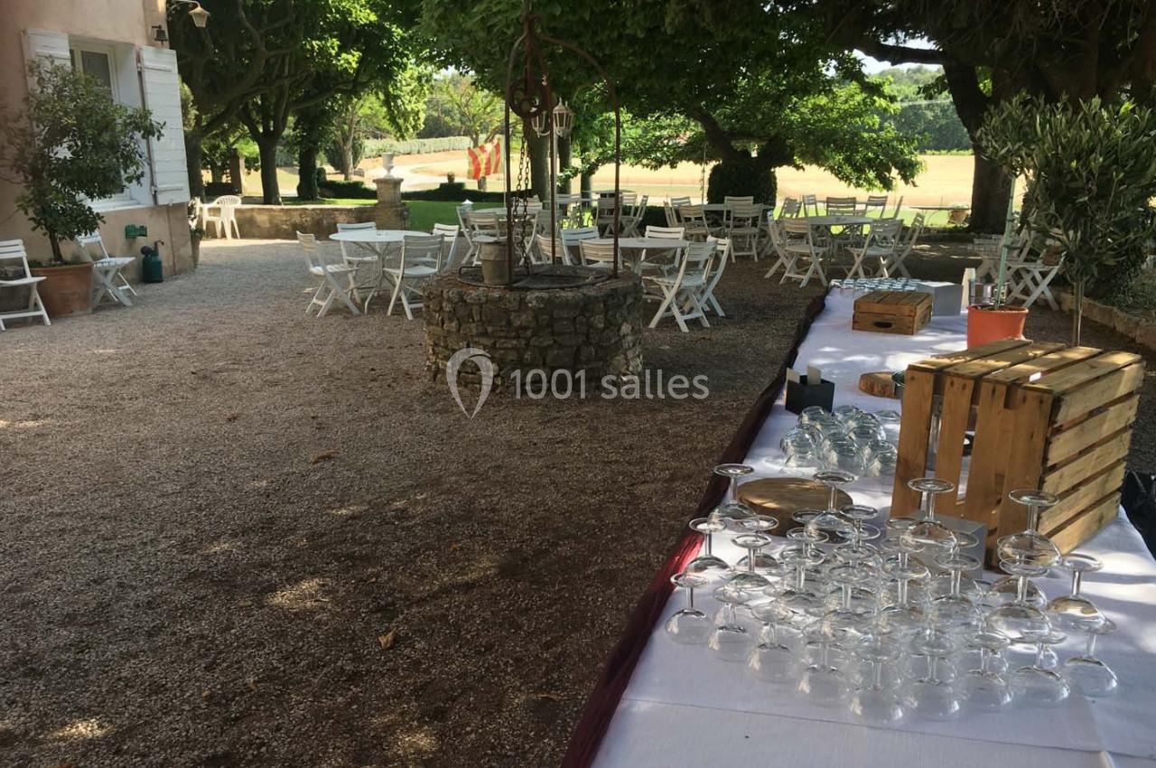 Tables et chaises blanches disposées en extérieur sous des arbres, avec un buffet et des verres alignés au premier plan.