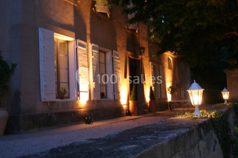 Façade d'une maison en soirée, éclairée par des lampes extérieures et entourée de végétation.