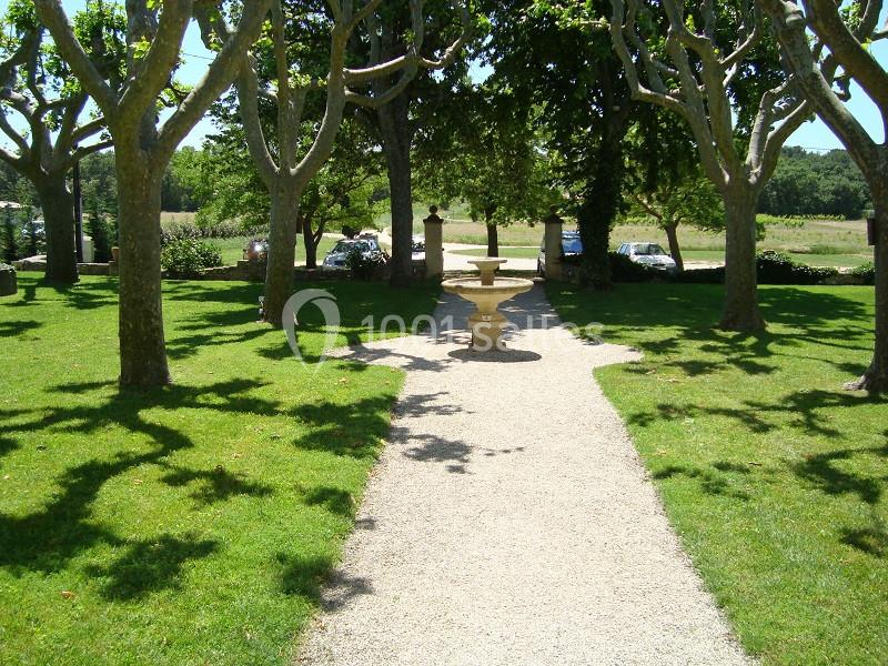 Allée bordée d'arbres menant à une fontaine en pierre, entourée de pelouse et d'un paysage champêtre.