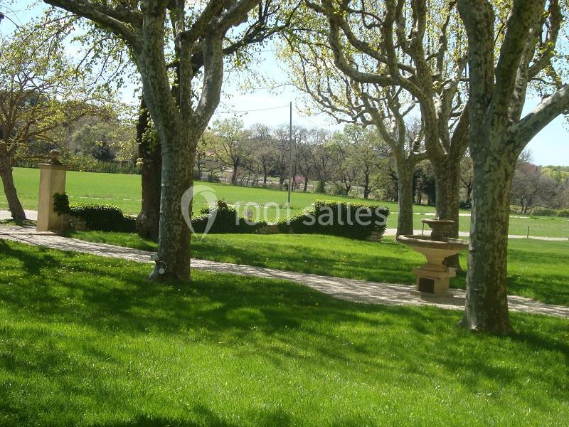 Pelouse verdoyante bordée d'arbres et d'une haie, avec un chemin pavé et une fontaine en pierre au premier plan.