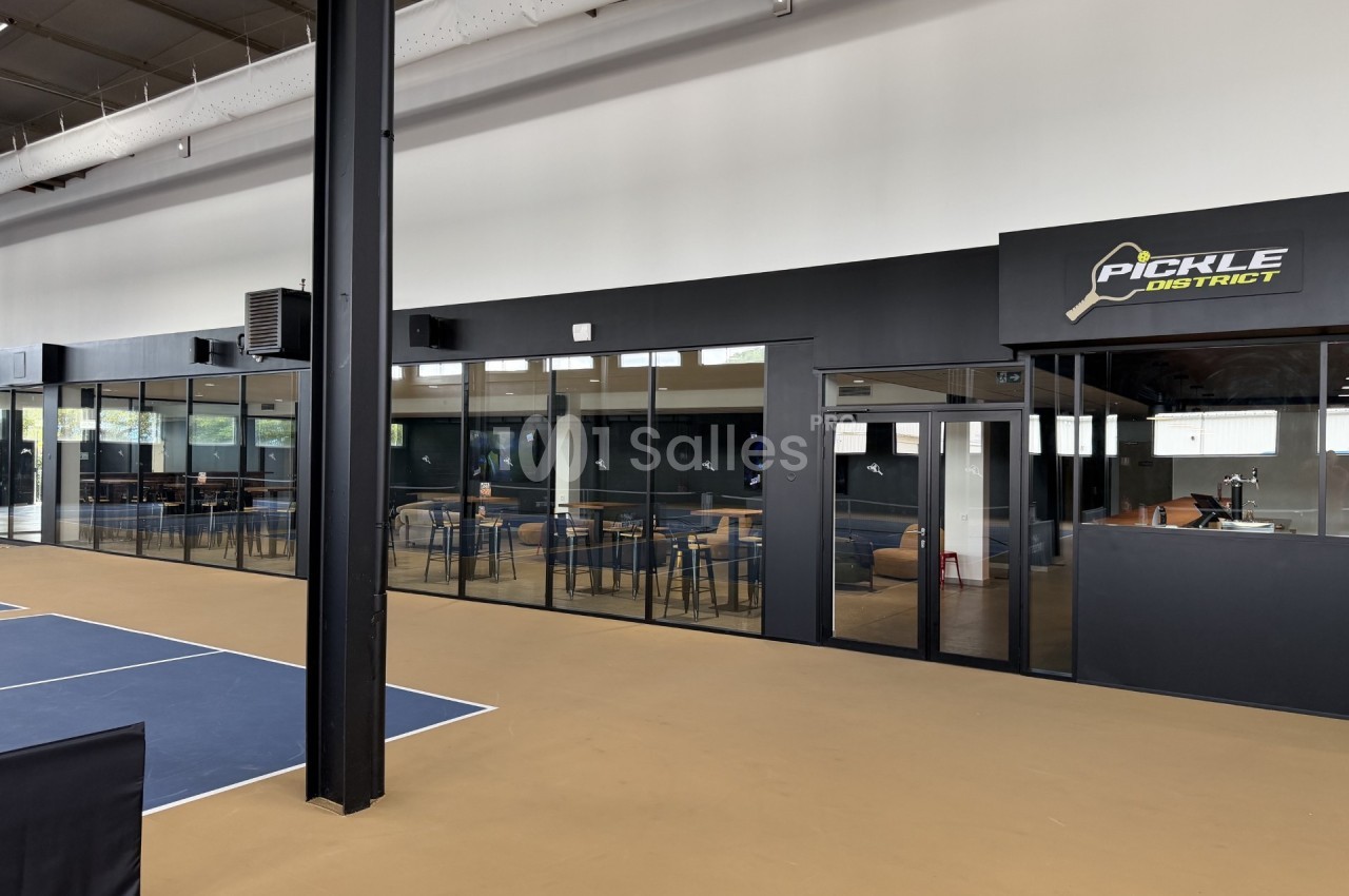 Grande salle sportive avec un terrain bleu délimité de blanc, à côté d'un espace bar vitré doté de hautes tables et de tabourets.