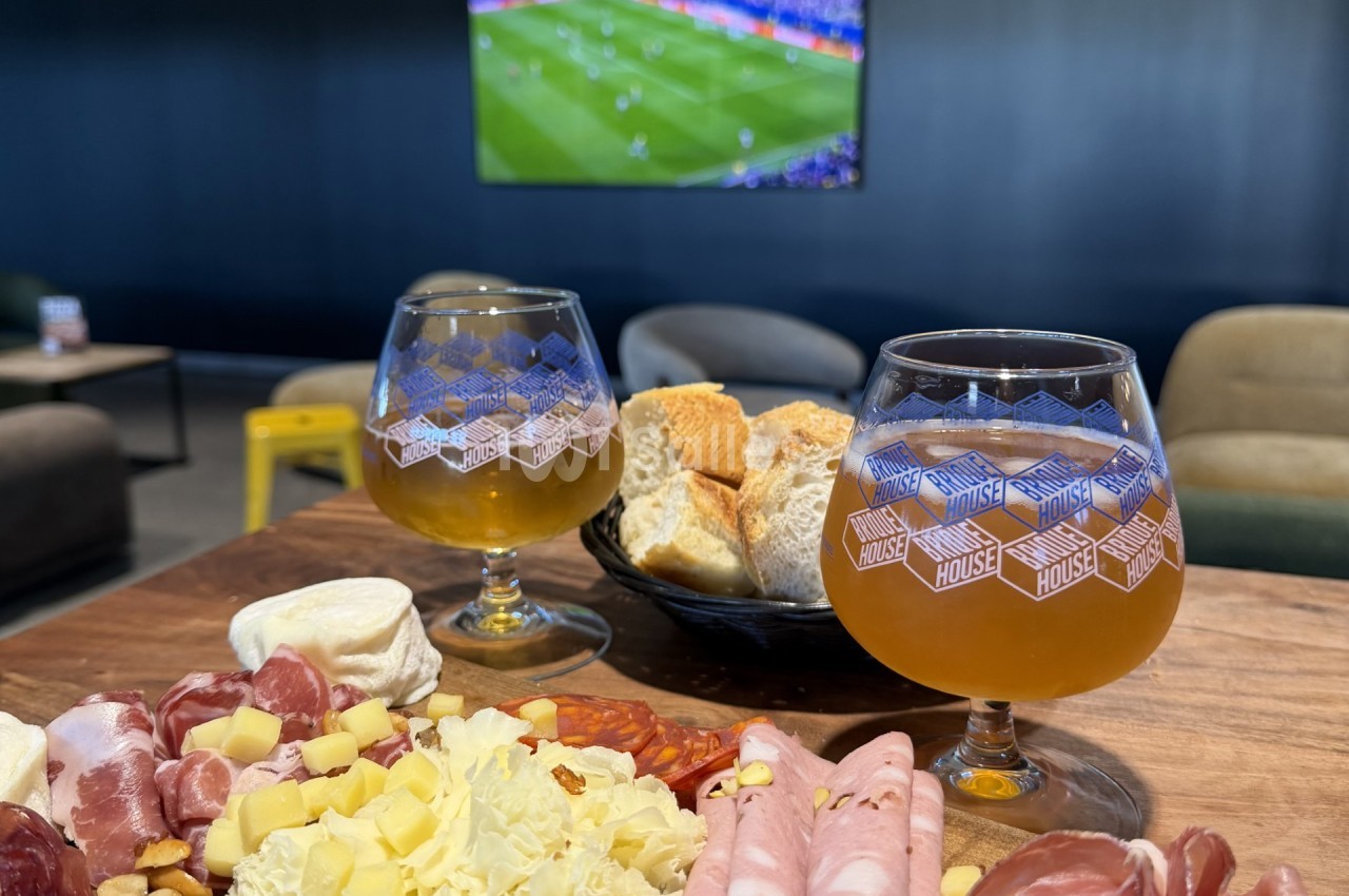 Deux verres de bière ambrée à motif « Brew House » sur une table en bois, devant une planche de charcuteries, de fromages et de pain, avec un écran de football en arrière-plan. Count: let me estimate length. "Deux verres de bière ambrée à motif « Brew