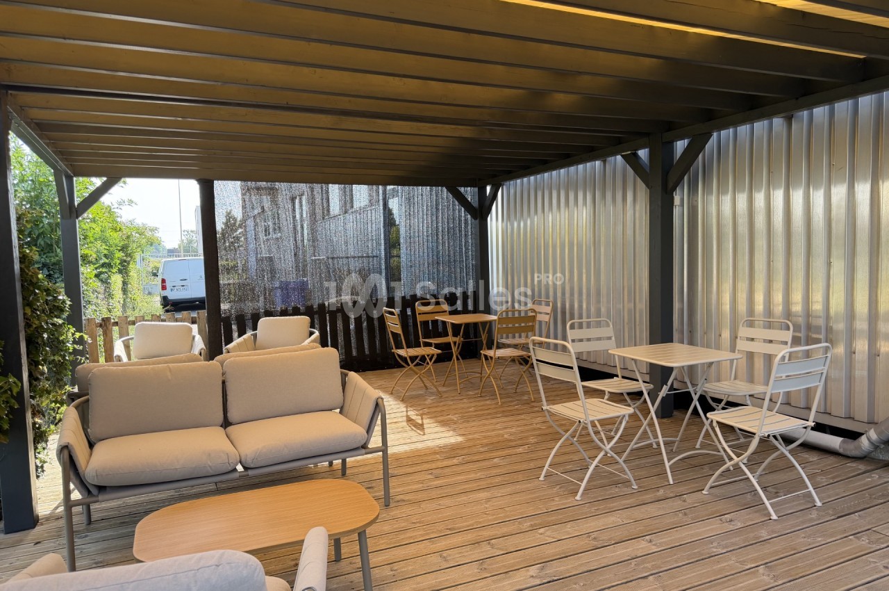 Terrasse en bois couverte par une pergola sombre, avec canapés à coussins beiges, tables basses et chaises pliantes blanches et dorées disposées sur le platelage. Let me check: under 125 characters? Count roughly: "Terrasse en bois couverte par une