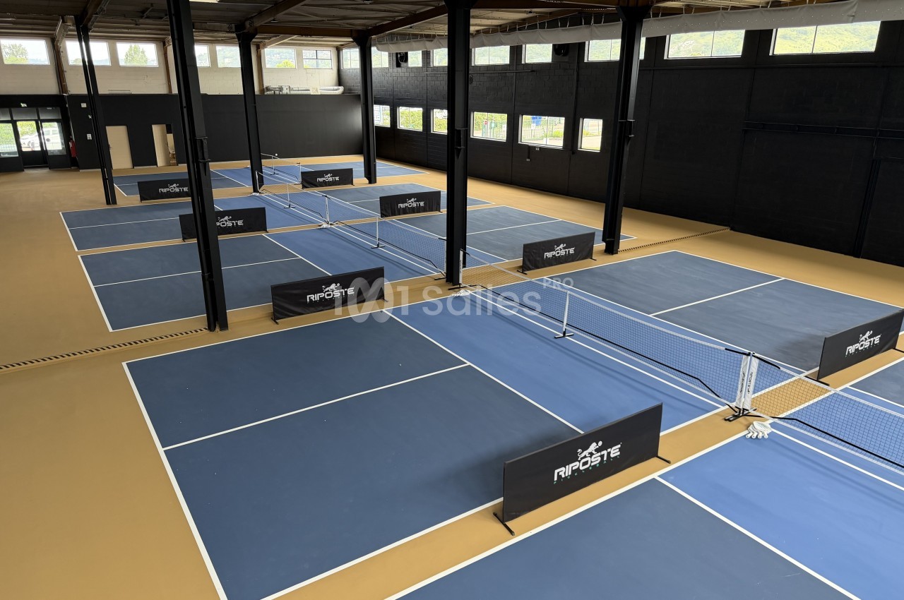 Hall couvert avec plusieurs terrains de pickleball bleus aux lignes blanches, séparés par des filets et des panneaux noirs, sous une charpente métallique.