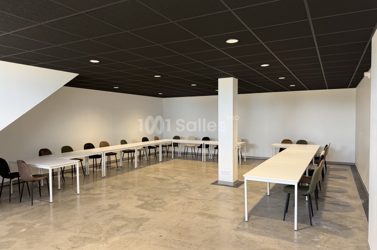 Salle vide à sol béton ciré, équipée de tables blanches disposées en U avec des chaises de couleurs variées, sous un plafond noir et un éclairage encastré. Let me verify length: roughly 150 characters, too long. Need ≤125. Salle au sol en béton ciré avec