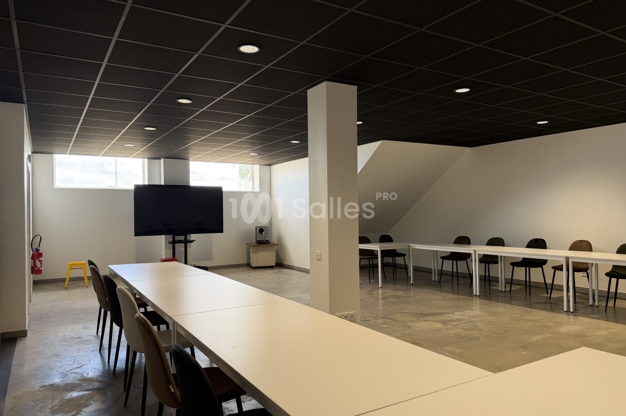 Salle de séminaire aux murs blancs et sol en béton ciré, avec tables blanches alignées, chaises variées et un écran TV fixé sur pied. Check: length ~150 characters? Let's count roughly: "Salle de séminaire aux murs blancs et sol en béton ciré, avec