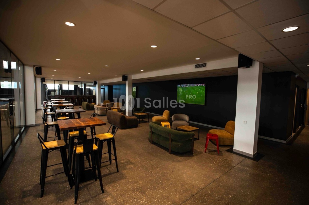 Salle intérieure aux murs sombres avec tables hautes en bois et tabourets noirs, canapés et fauteuils colorés, et un écran de retransmission sportive fixé au mur.
