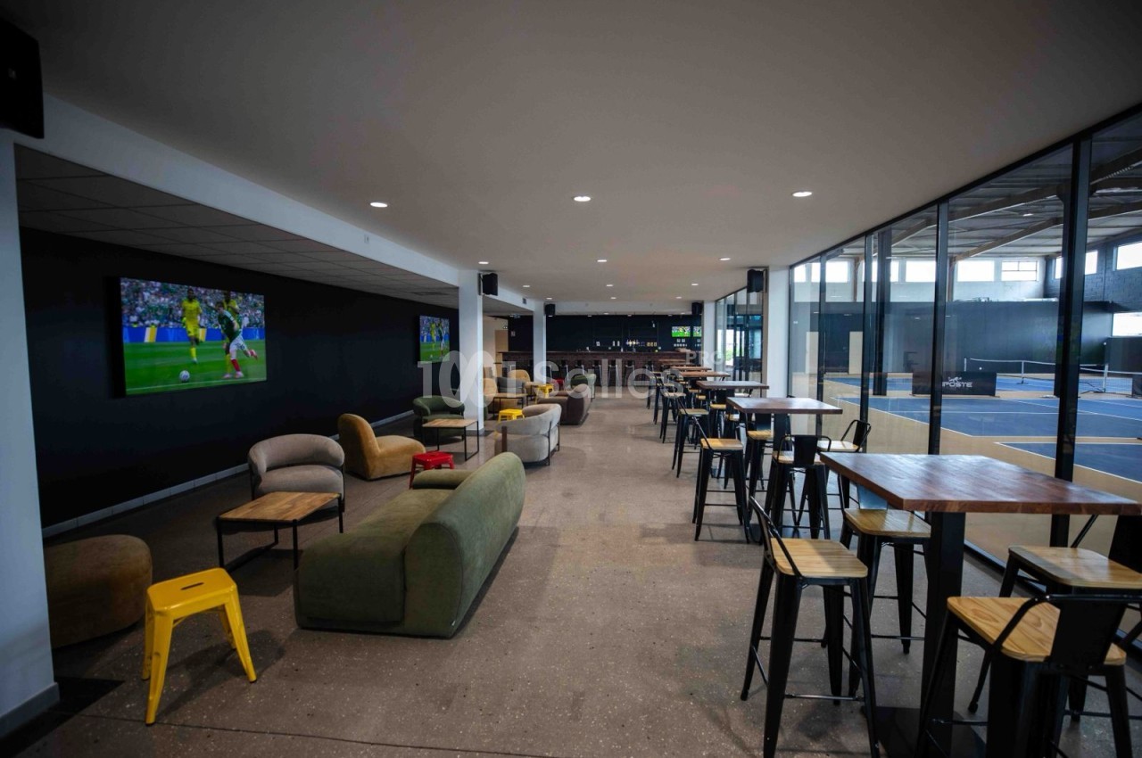 Espace lounge avec canapés, fauteuils et tables hautes, téléviseur mural et baies vitrées donnant sur des courts couverts.