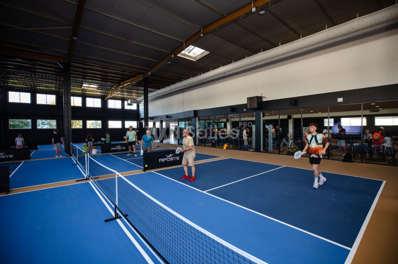 Intérieur d'un grand hall avec plusieurs terrains de pickleball bleus délimités par des filets, où des joueurs s'exercent en raquette sous une charpente métallique. Let me check constraints: one sentence, under 125 chars? That sentence is about 170