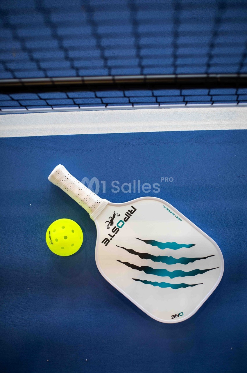Raquette de pickleball blanche à motifs bleu-vert en griffures posée sur un court bleu, à côté d'une balle jaune fluo, filet en arrière-plan.