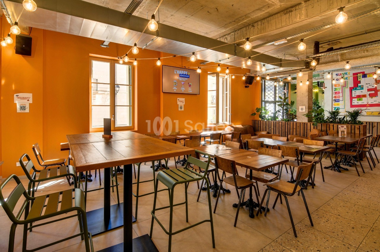Salle de restaurant aux murs orangés, avec tables en bois et chaises métal vertes, éclairée par des guirlandes de guinguettes suspendues sous une structure béton apparente. Wait, I must respect the rules: no "guinguettes" invention? Guirlandes d'ampoules