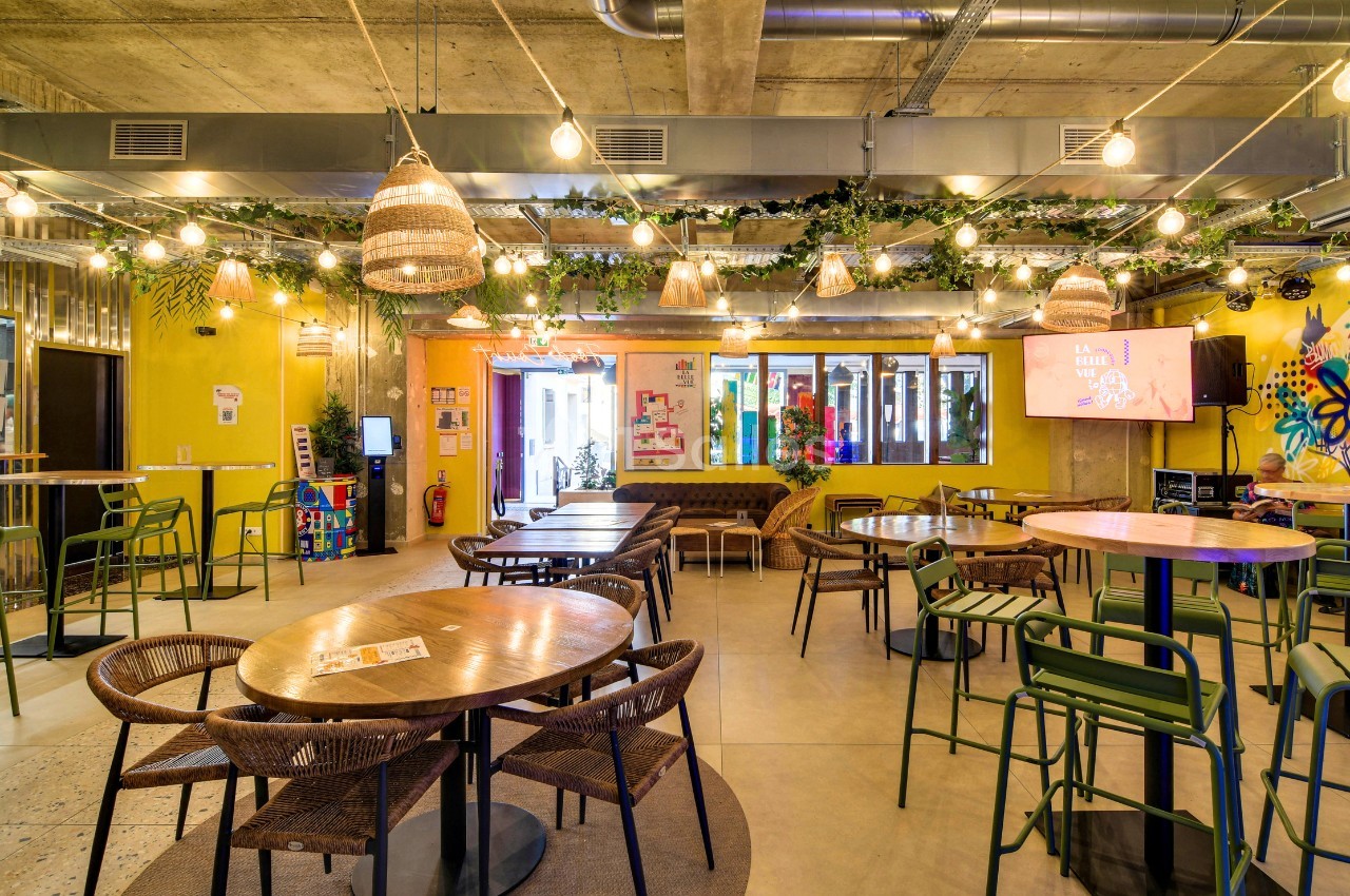 Salle lumineuse aux murs jaunes, avec tables rondes en bois, chaises en osier et mange-debout verts, sous des guirlandes d'ampoules et des plantes suspendues.
