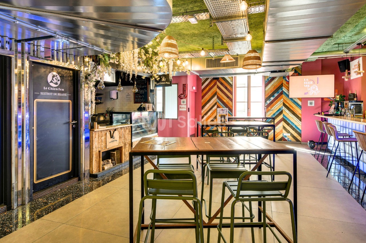 Salle de restauration aux tables hautes en bois et chaises vertes, murs roses et lambris colorés, comptoir de bar et petit stand de comptoir visibles au fond.
