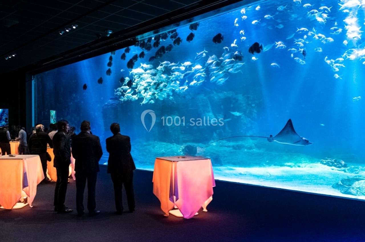 Des personnes debout près de tables éclairées regardent un grand aquarium avec des poissons et une raie nageant.