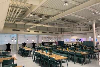 Salle lumineuse avec des tables en bois, des stands d'exposition et des personnes observant ou manipulant des objets.