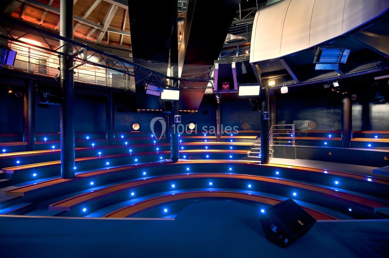 Salle de conférence circulaire avec gradins éclairés par des lumières bleues et plafond industriel.