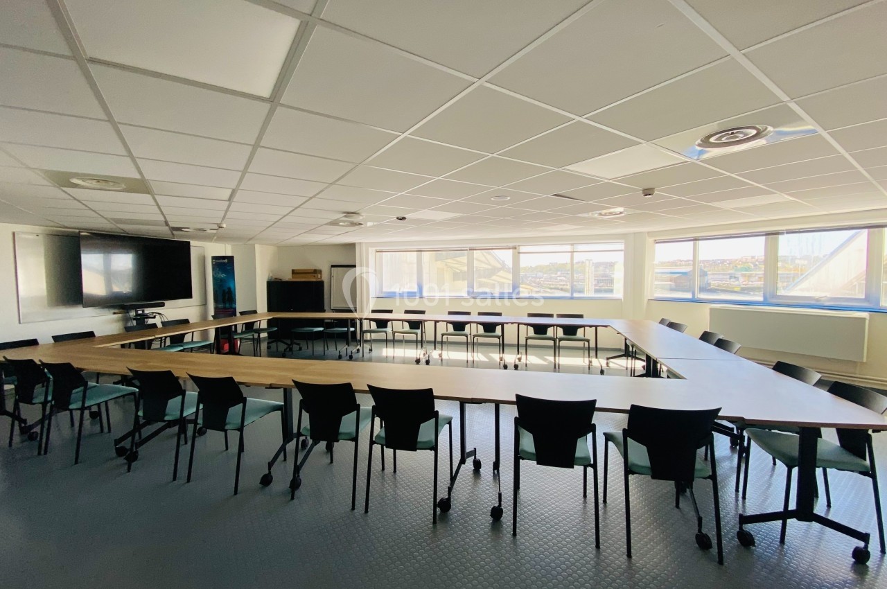 Salle de réunion lumineuse avec une grande table en U entourée de chaises noires, écran et fenêtres en arrière-plan.